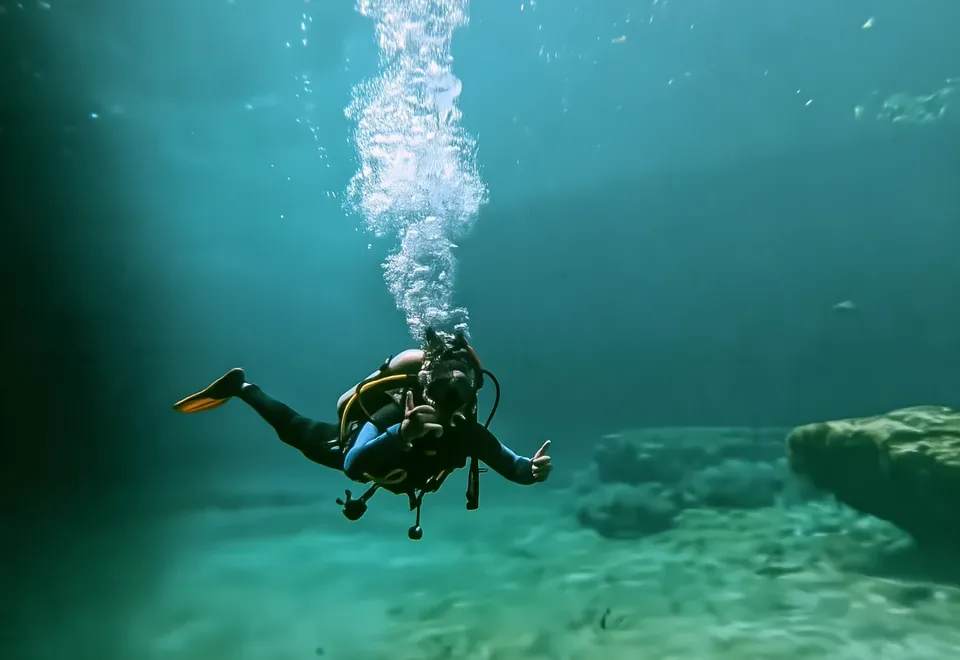 A diver giving a thumbs-up in the turquoise water of a Tulum cenote, a column of bubbles rising above