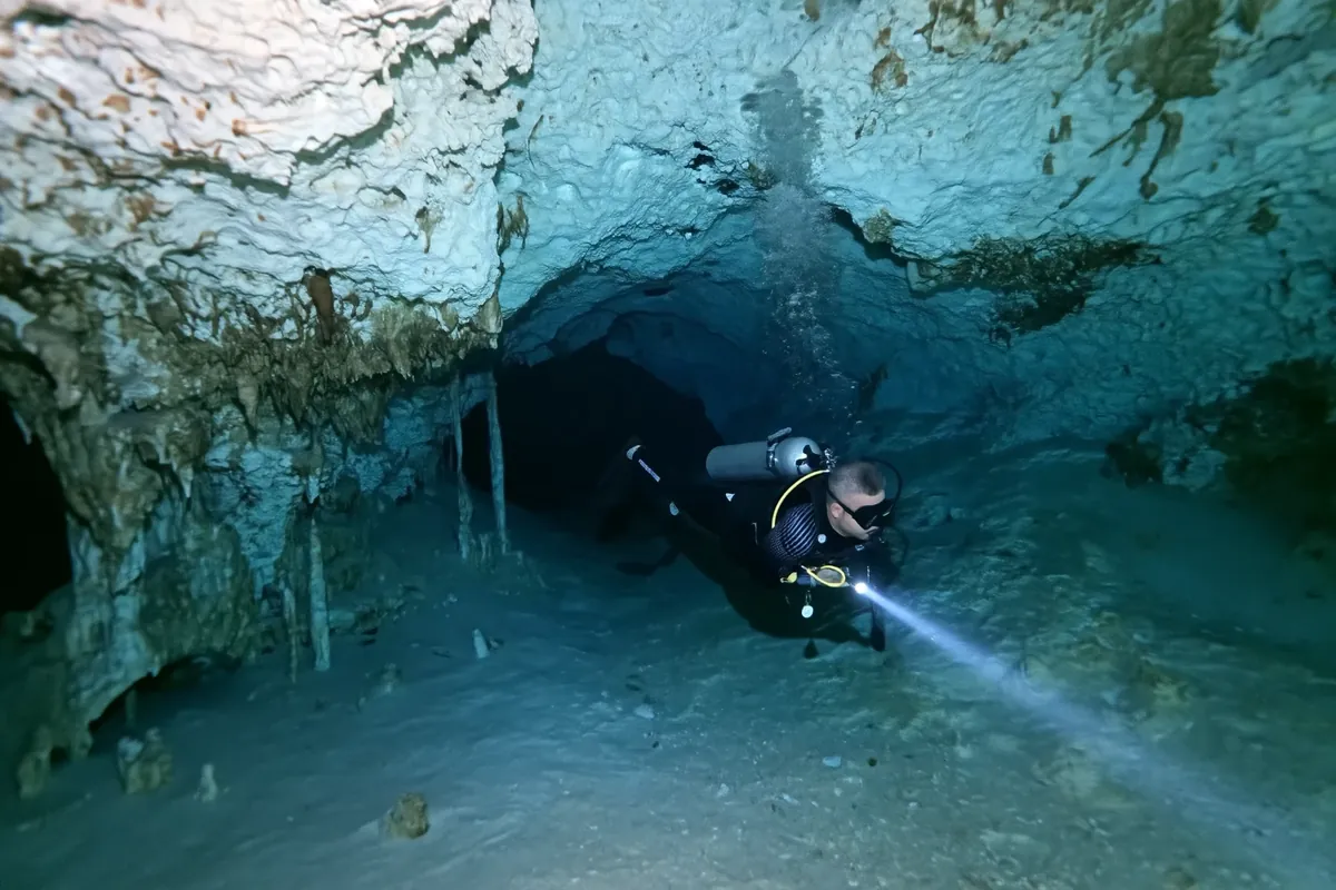 A diver with a torch lit swimming through a pale rock gallery in Cenote Tak Be Luum